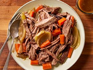 Overhead view of a plate of pot roast with carrots and onions and a fork and spoon all next to a cup of gravy on a wooden tabletop