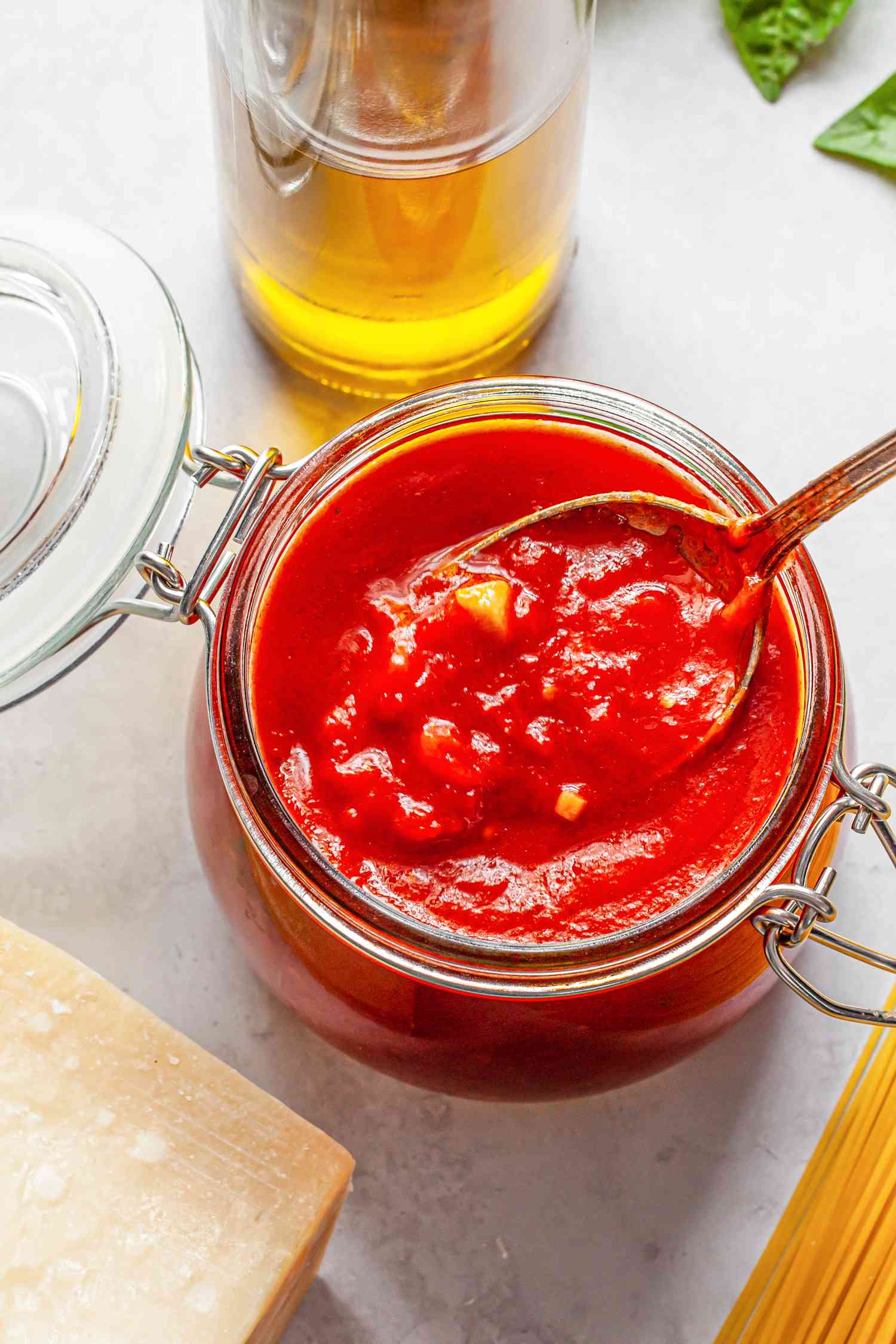 Jar of Homemade Marinara Sauce With a Spoon, and in the Surroundings, a Bottle of Olive Oil, Fresh Basil Sprigs, Dry Pasta, and a Block of Parmesan