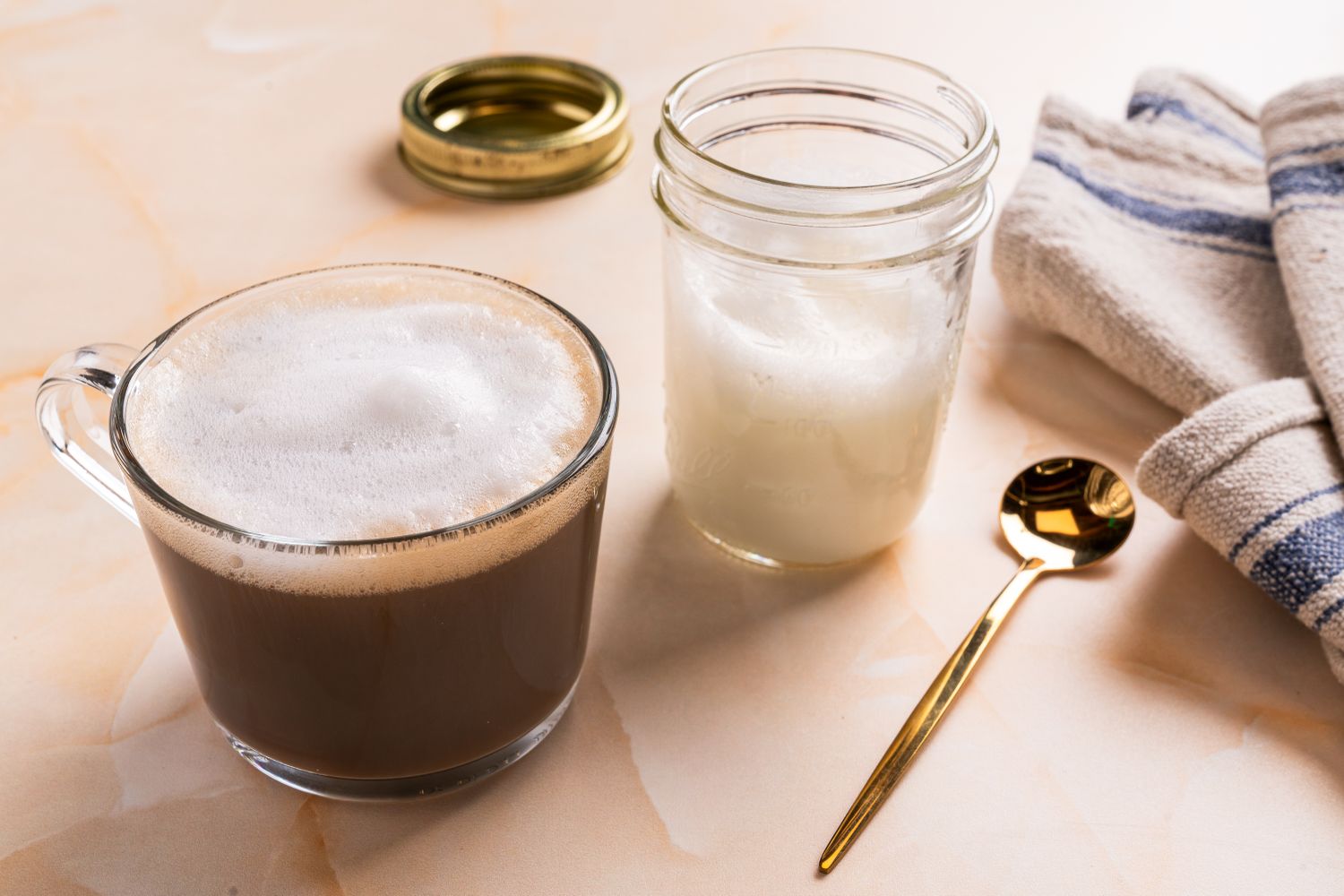 Cup of coffee with frothy milk accompanied by a jar of milk froth a golden spoon and a striped cloth napkin