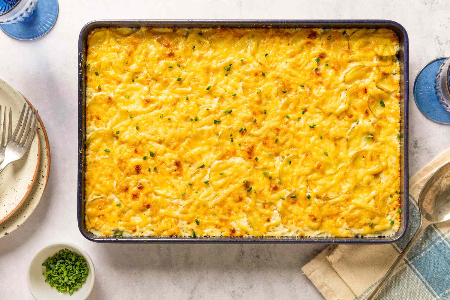 Overhead view of a 9x13 sheet pan of Sheet Pan Au Gratin Potatoes recipe on a countertop with forks, a serving spoon, glasses and a small bowl of chives