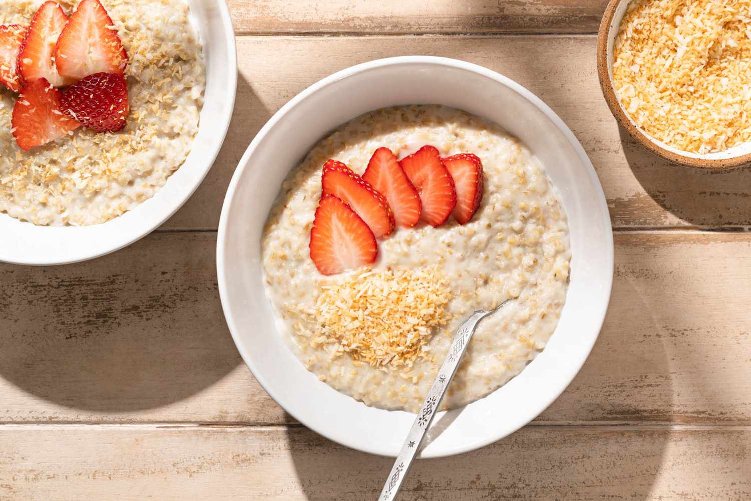 Bowl of oatmeal topped with sliced strawberries and shredded coconut, spoon resting on the bowl, additional oatmeal and toppings nearby.