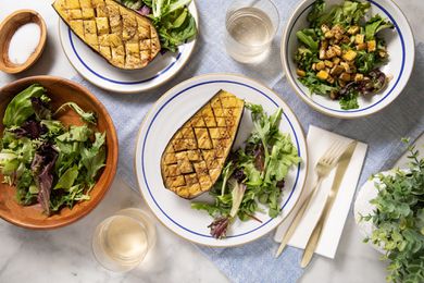 Table Setting (L to R): Small Bowl of Salt, Bowl of Salad Greens, Two Plates With Halved Eggplants and a Serving of Side Salad, Two Glasses, a Napkin With a Fork and Knife, a Bowl of Salad Greens Topped With Cubed Eggplant, and a Small Indoor Plant