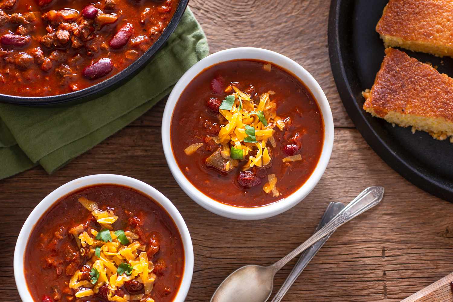 Overhead view of two white serving bowls of chili on a wooden tabletop next to two spoons, a large bowl of chili and a plate of corn bread