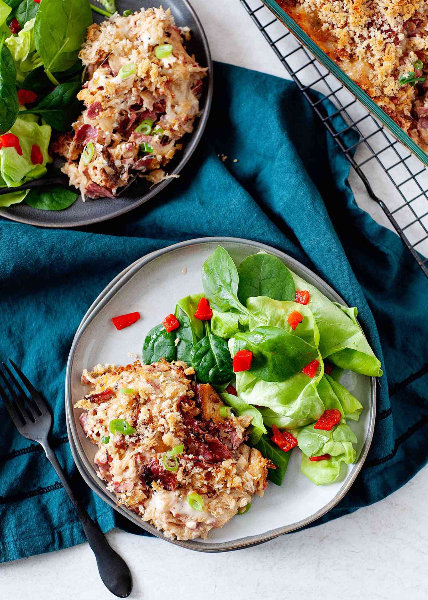 Overhead vertical view of a ceramic plate with a scoop of easy reuben casserole and a side salad with diced red peppers. A dark teal linen is under the plate and a second plate mirroring the first is behind it. A black fork is to the left and a cooling rack with a partial view of the casserole dish is in the upper right hand corner.