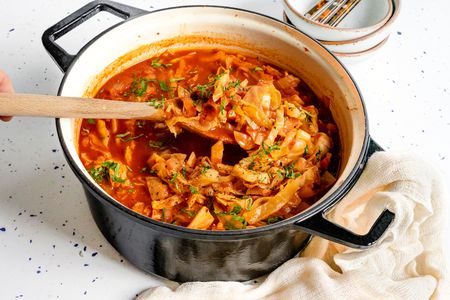 Sweet and Sour Cabbage Soup in a Large Pot with Some Raised Up Using a Wooden Spoon, and Surrounding the Pot, a Kitchen Linen and a Stack of Bowls with Utensils
