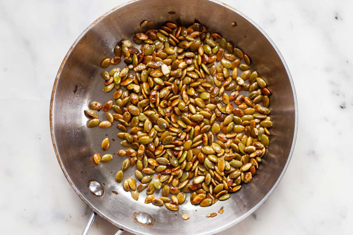 Silver saute pan on a marble background with toasted pumpkin seeds inside.