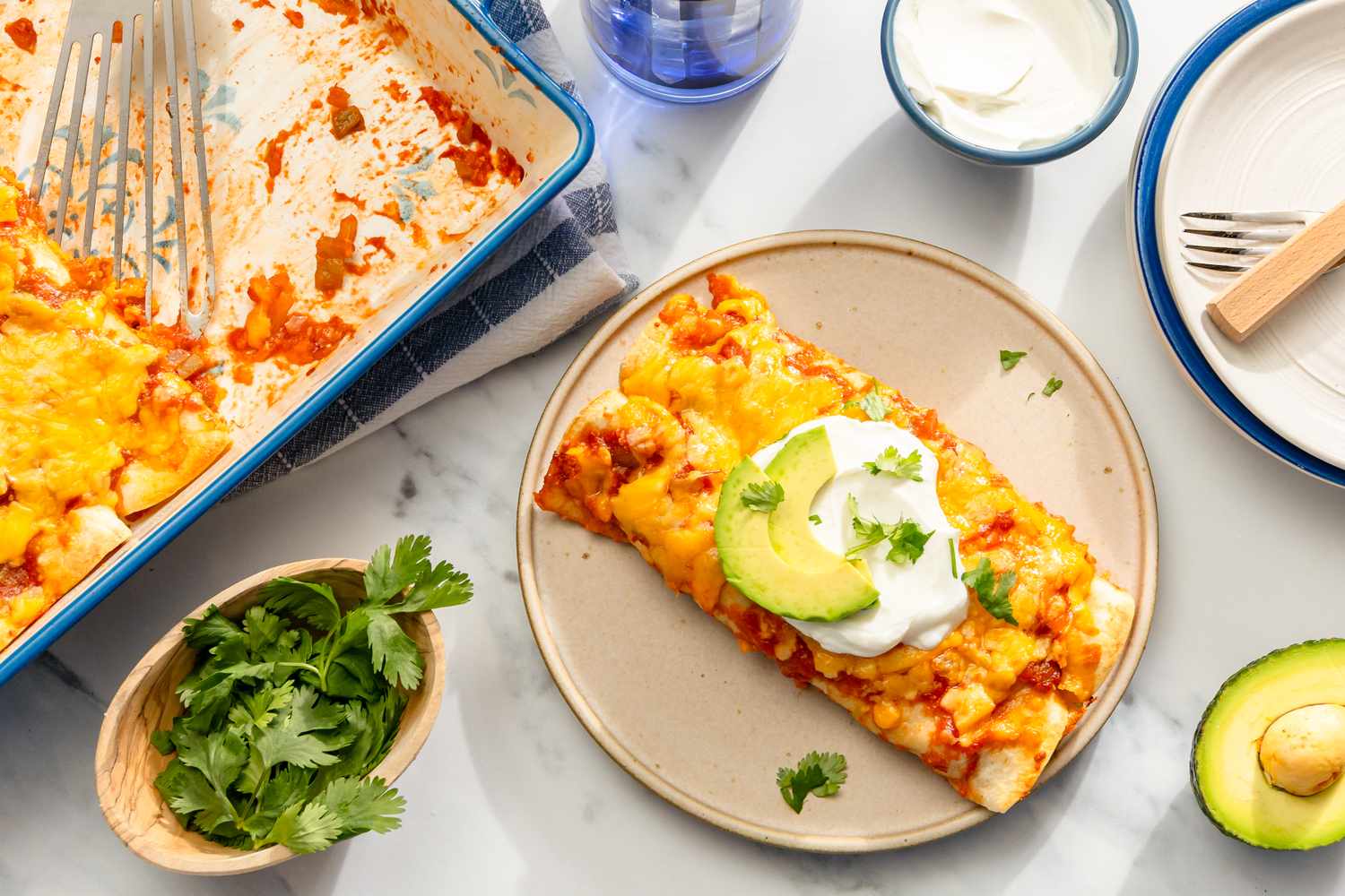 A plate with a chicken and spinach tortilla bake topped with avocado and sour cream served beside a casserole dish and garnishes