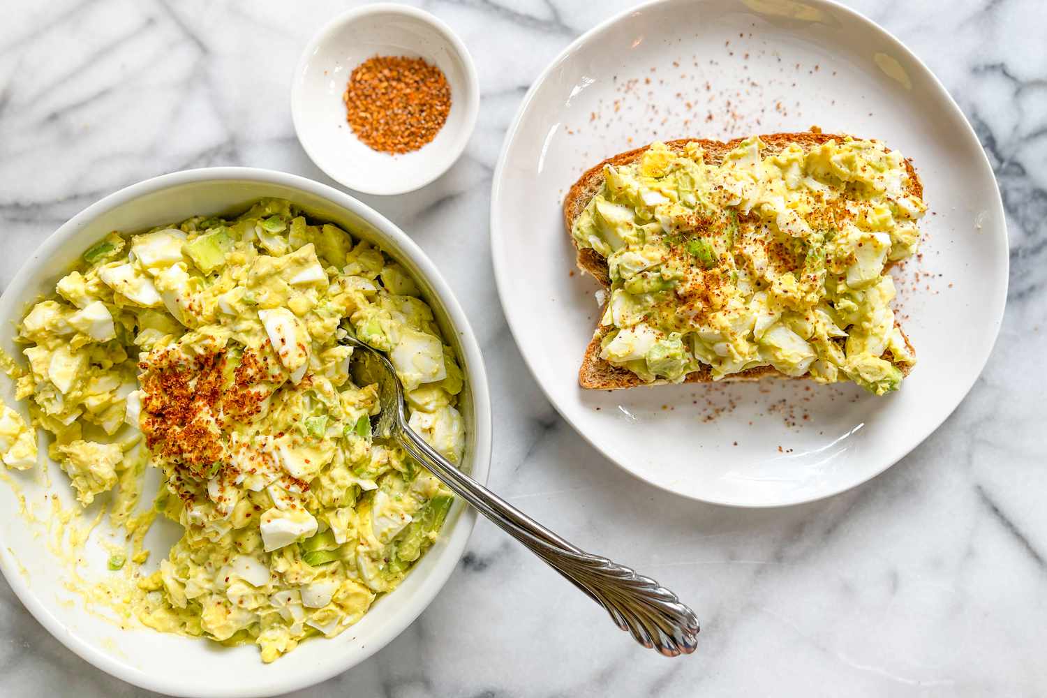Tajin egg salad in a bowl with a spoon next to a plate with egg salad-topped slice of bread and a bowl with more Tajin 