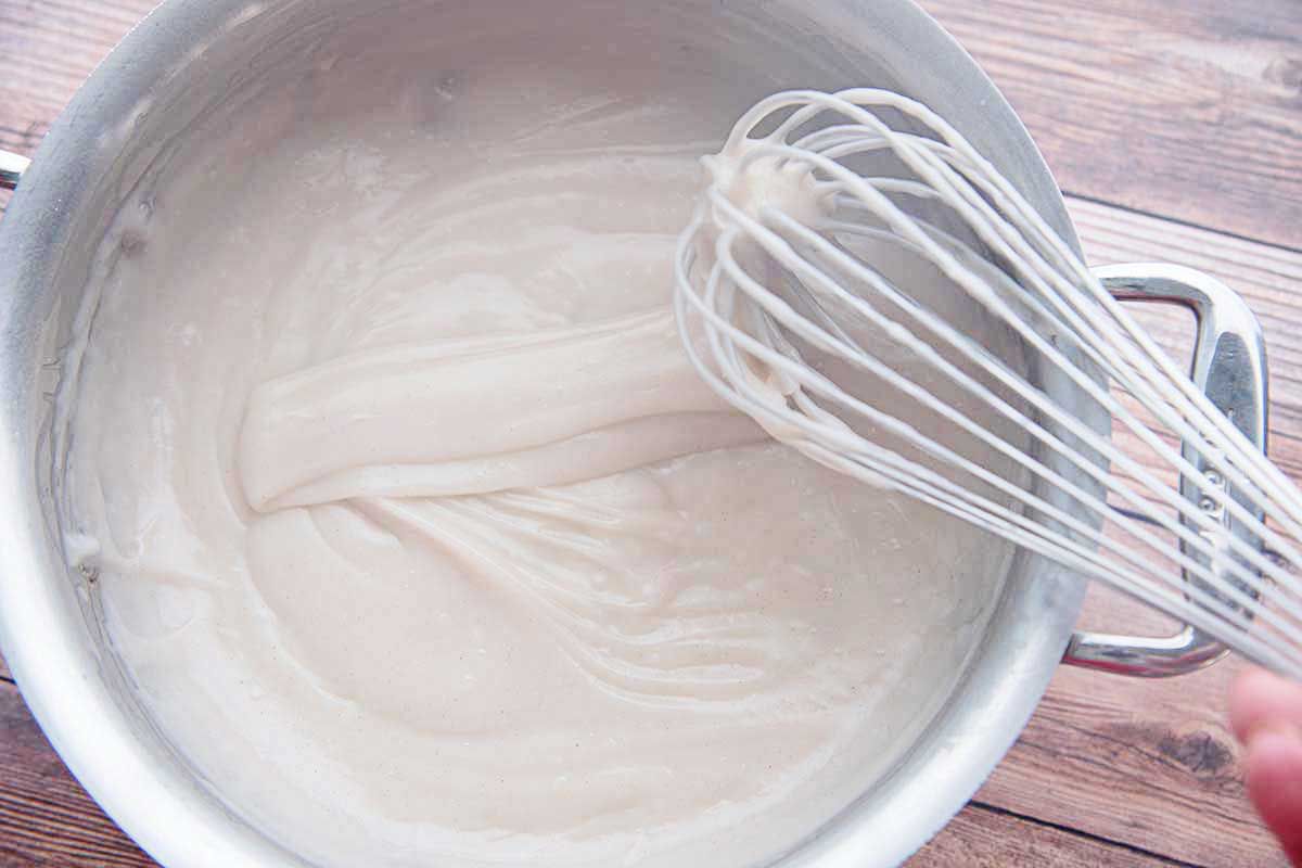 A metal pan has easy templeque inside and a whisk is coated in the mixture and lifted above the pan.