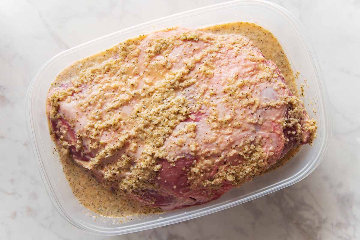 Cuban-style pork shoulder submerged in marinade in a clear plastic container. The container sits on a marble background.