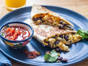 Side view of two simple breakfast quesadillas on a blue plate with a bowl of salsa to the left and a glass of orange juice above. The scrambled eggs, black beans and cilantro are visible inside the quesadilla.