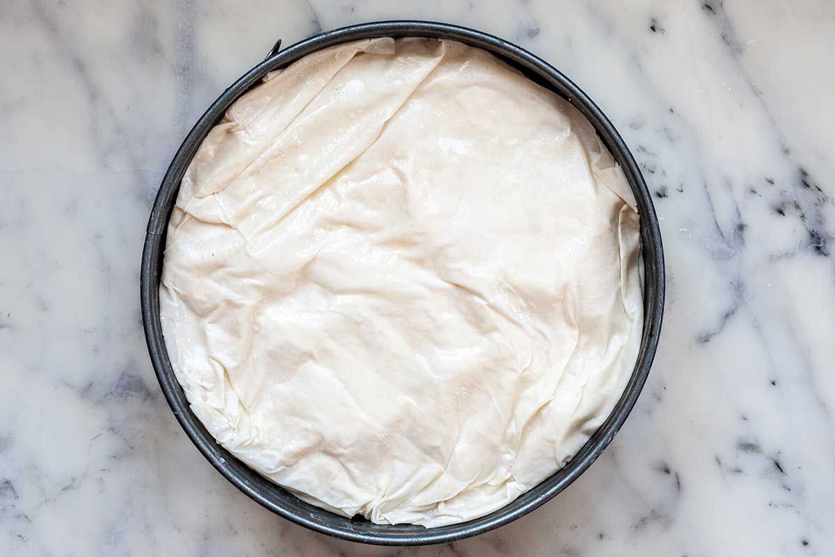 A cake pan is on a marble countertop. Unbaked phyllo dough is covering the top.