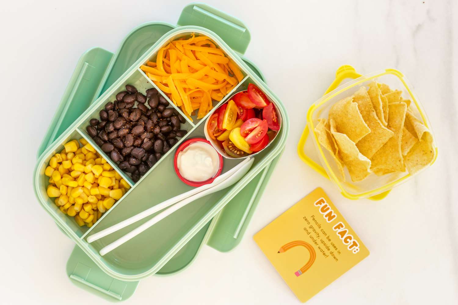Overhead view of a bento-style lunch box with corn, black beans, shredded cheese, tomatoes and sour cream and plasticware on a marble countertop next to a small container of tortilla chips