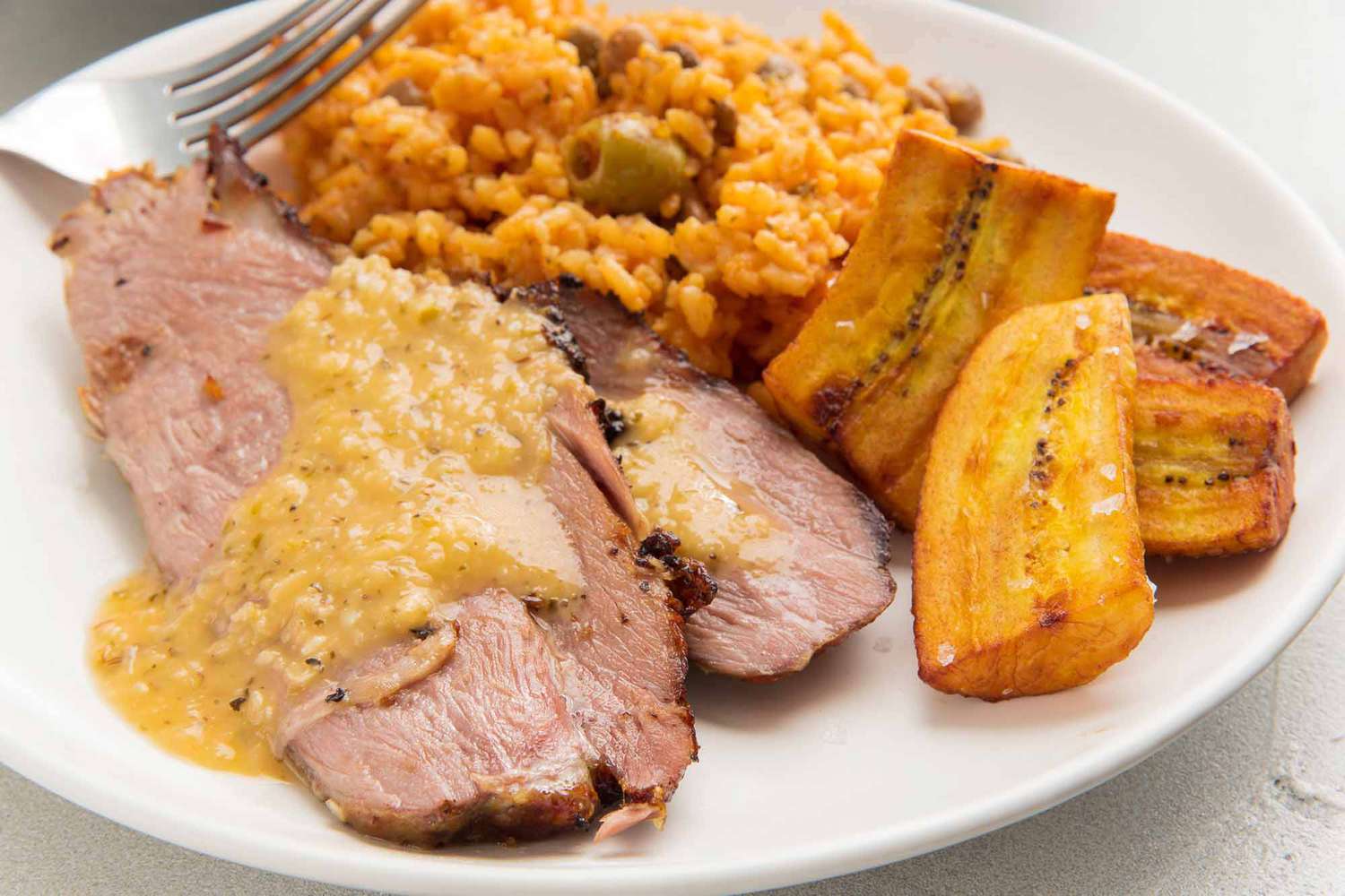 A white plate of sliced cuban style pork shoulder with marinade spooned on top. Fried plantains and red rice are on the plate as well.