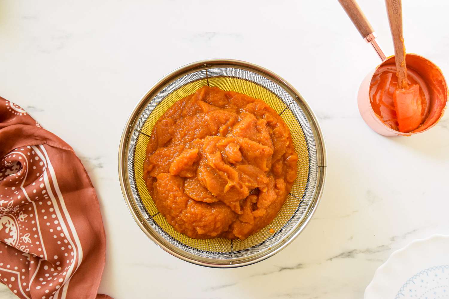 Pumpkin pie filling in a fine mesh strainer next to a brown kitchen linen and a measuring cup with a small spatula for 2-ingredient pumpkin mousse recipe