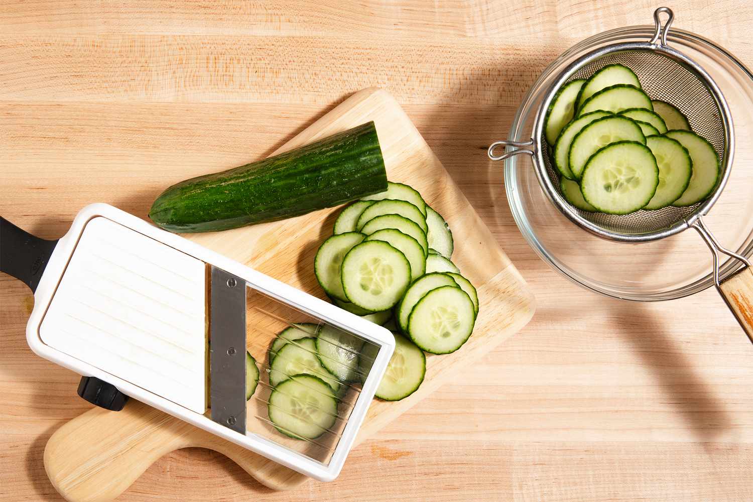 Thinly sliced cucumber on a cutting board with a mandoline slicer and a bowl