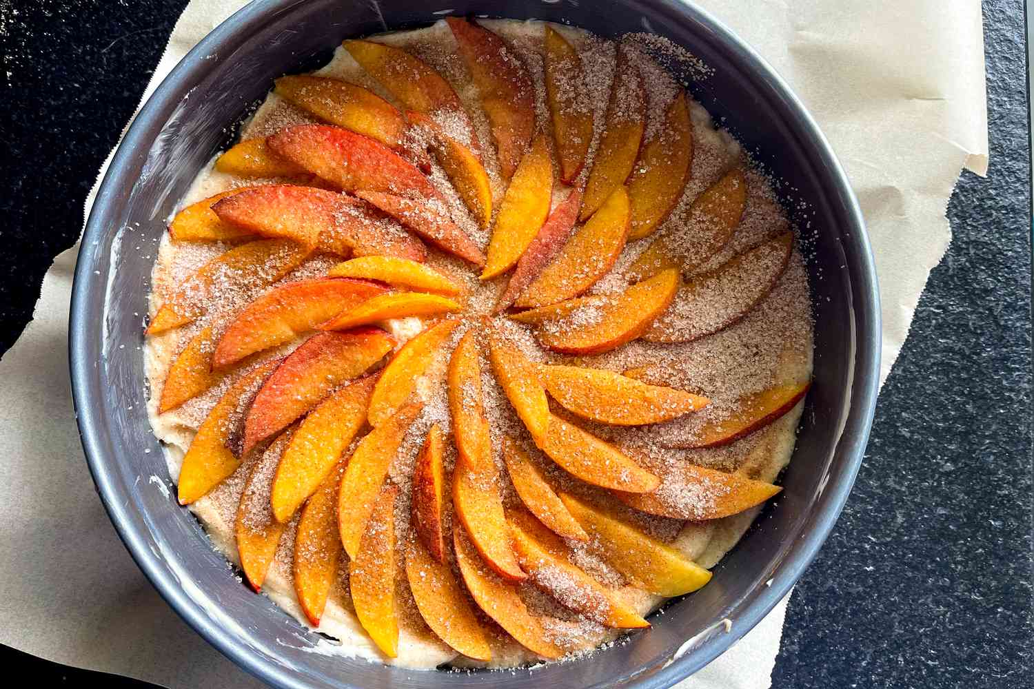 A baking pan with a cake being prepared, topped with sliced fruit arranged in a spiral pattern and dusted with sugar and spices