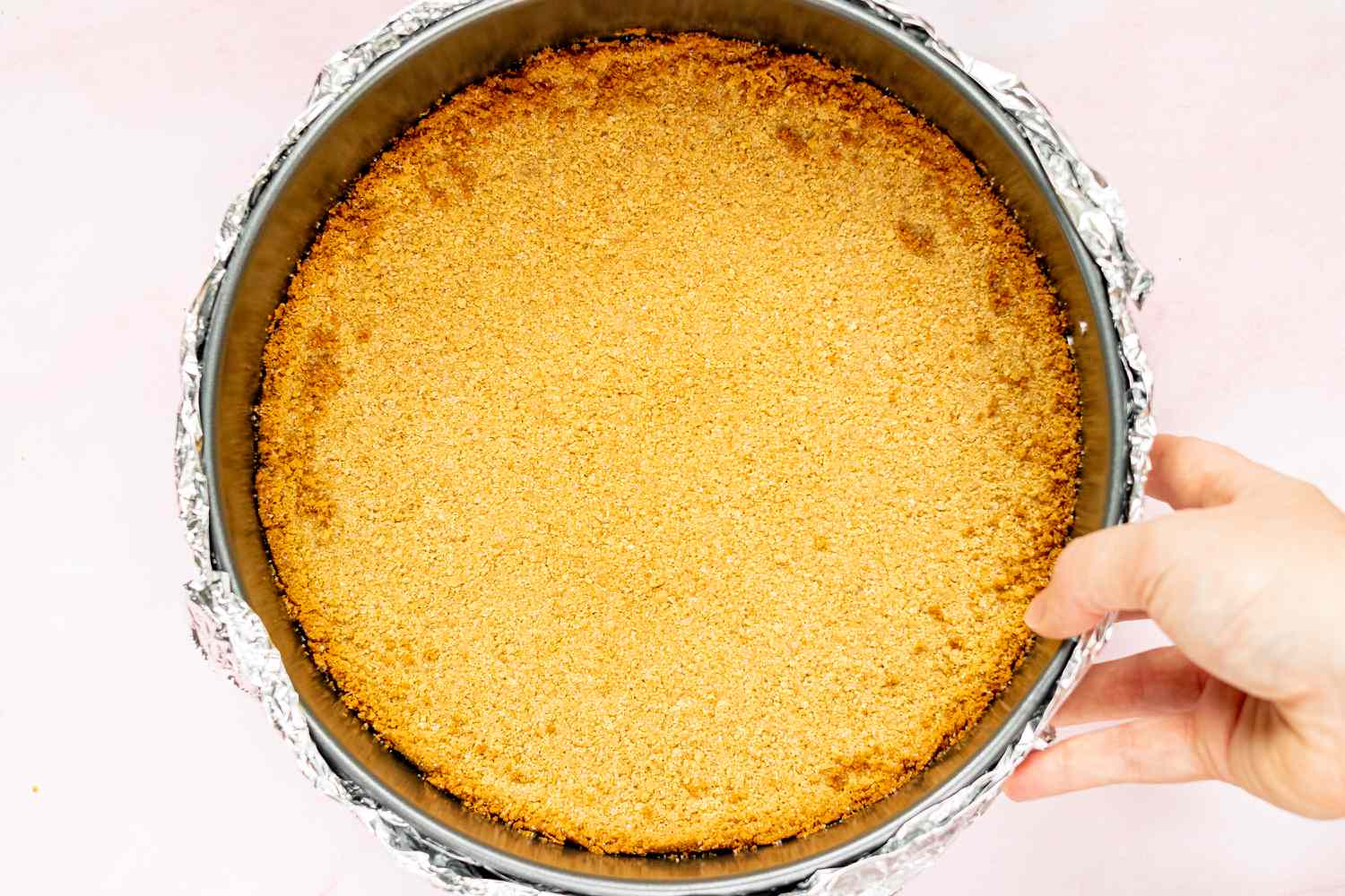 A hand holding the foil-wrapped springform pan with the crust inside 