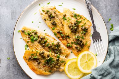 Sole piccata on white plate with a fork and slices of lemon, garnished with chopped parsley