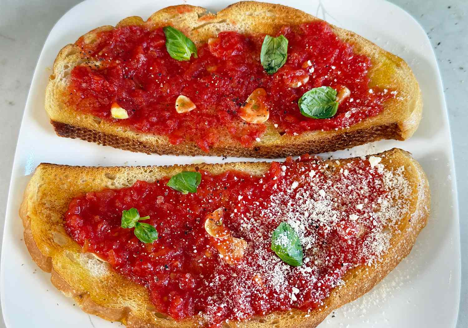 Two slices of toasted bread topped with crushed tomatoes basil leaves and garlic one slice sprinkled with grated cheese