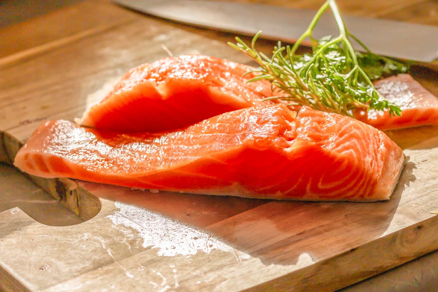 Angled view of two raw salmon fillets on a wooden cutting board topped with herbs and a large knife in the background
