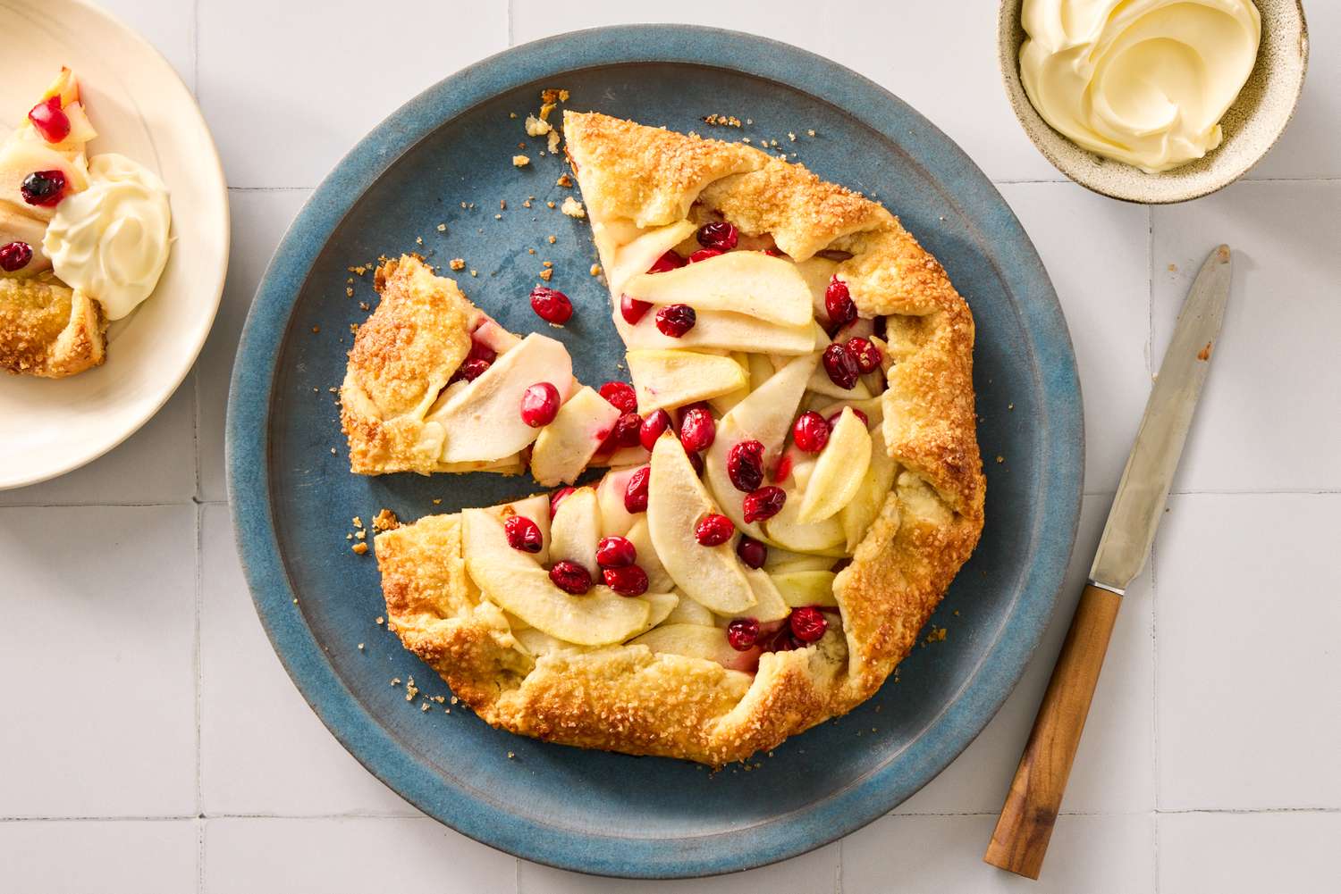 A pear and cranberry galette on a plate with a slice removed next to a serving of whipped cream
