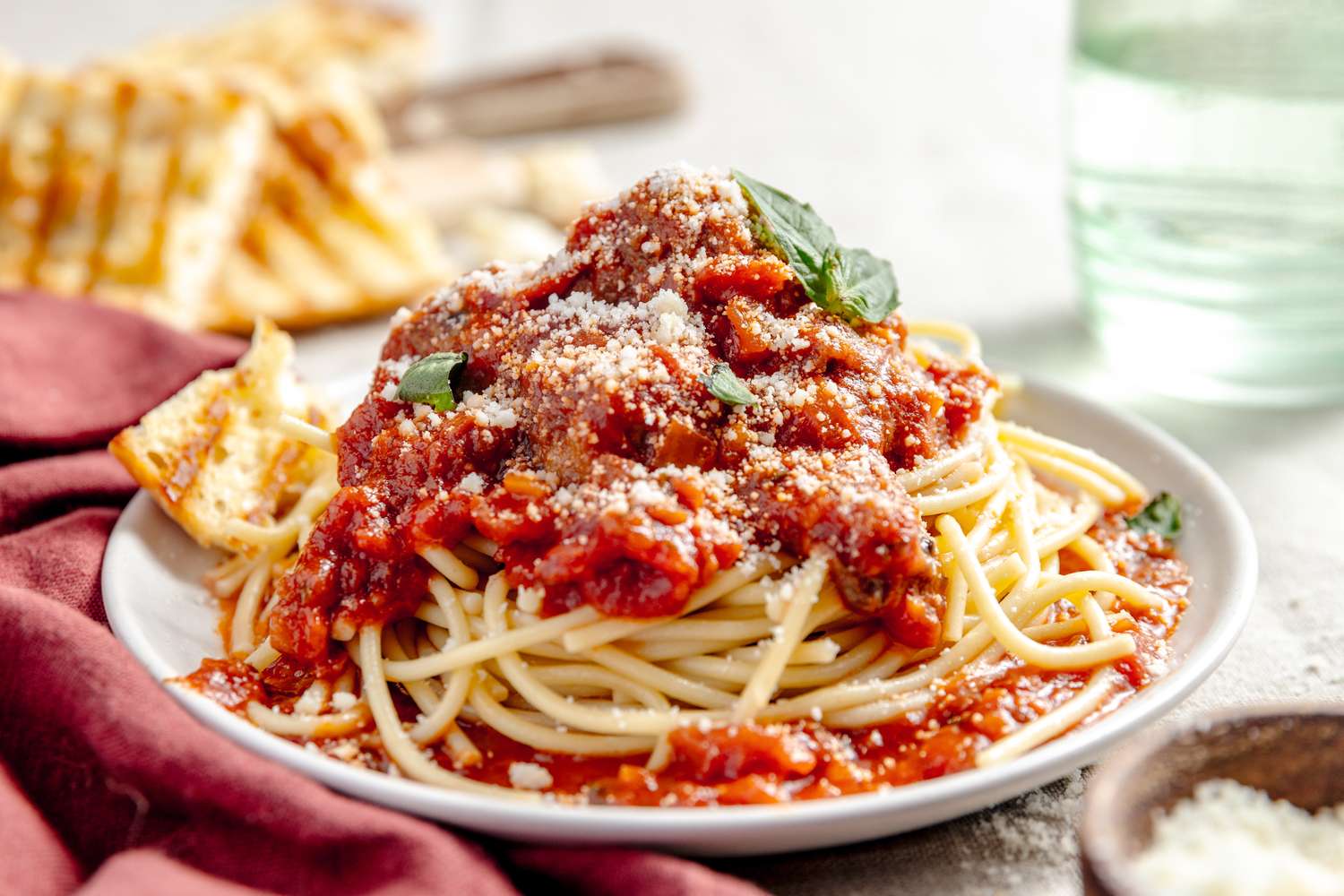 Plate of Spaghetti and Meatballs Topped with Basil, and in the Surroundings, a Glass of Water, Toasted Bread, a Burgundy Table Napkin, and a Bowl of Parmesan
