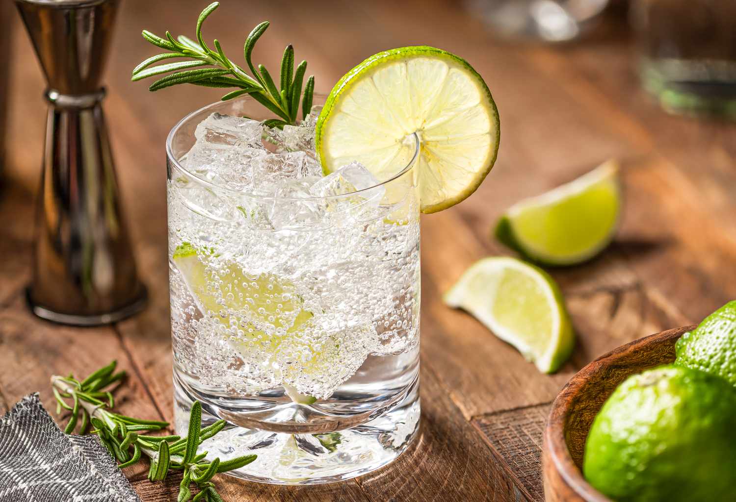 Glass of gin and tonic with lime garnished with a sprig of rosemary on a wooden table