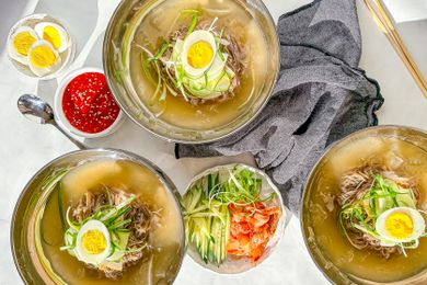 Three Bowls of Mul Naengmyeon Surrounded By Bowls of Toppings (One Bowl With Sliced Cucumbers, Kimchi, and Julienned Green Onions, Another With Spicy Gochujang Sauce, and a Bowl of Halved Hard Boiled Eggs), a Grey Kitchen Towel, and Utensils (a Spoon and Pair of Chopsticks)
