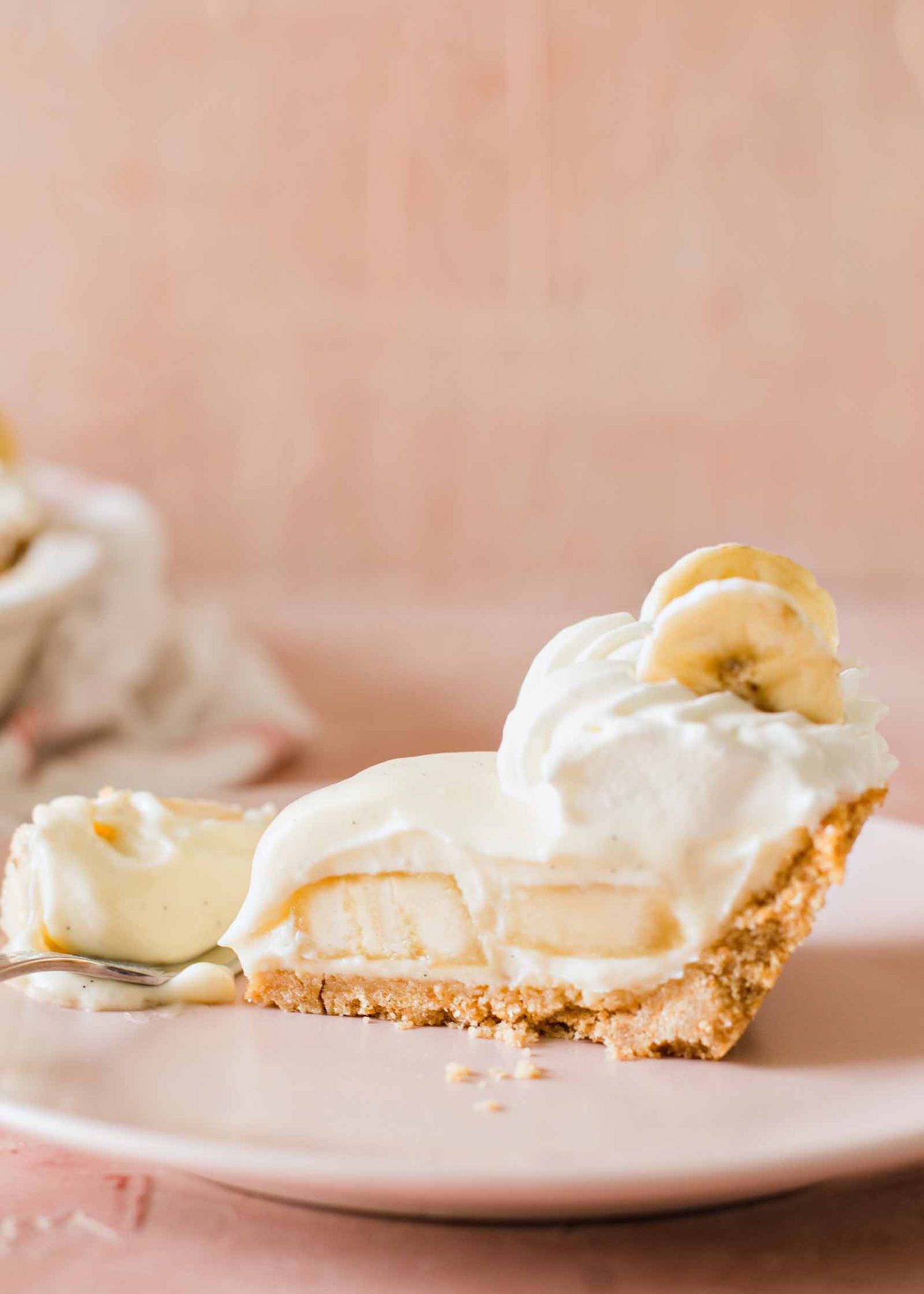 Slice of banana cream pie with whipped cream on top and banana slices set against a pink background.