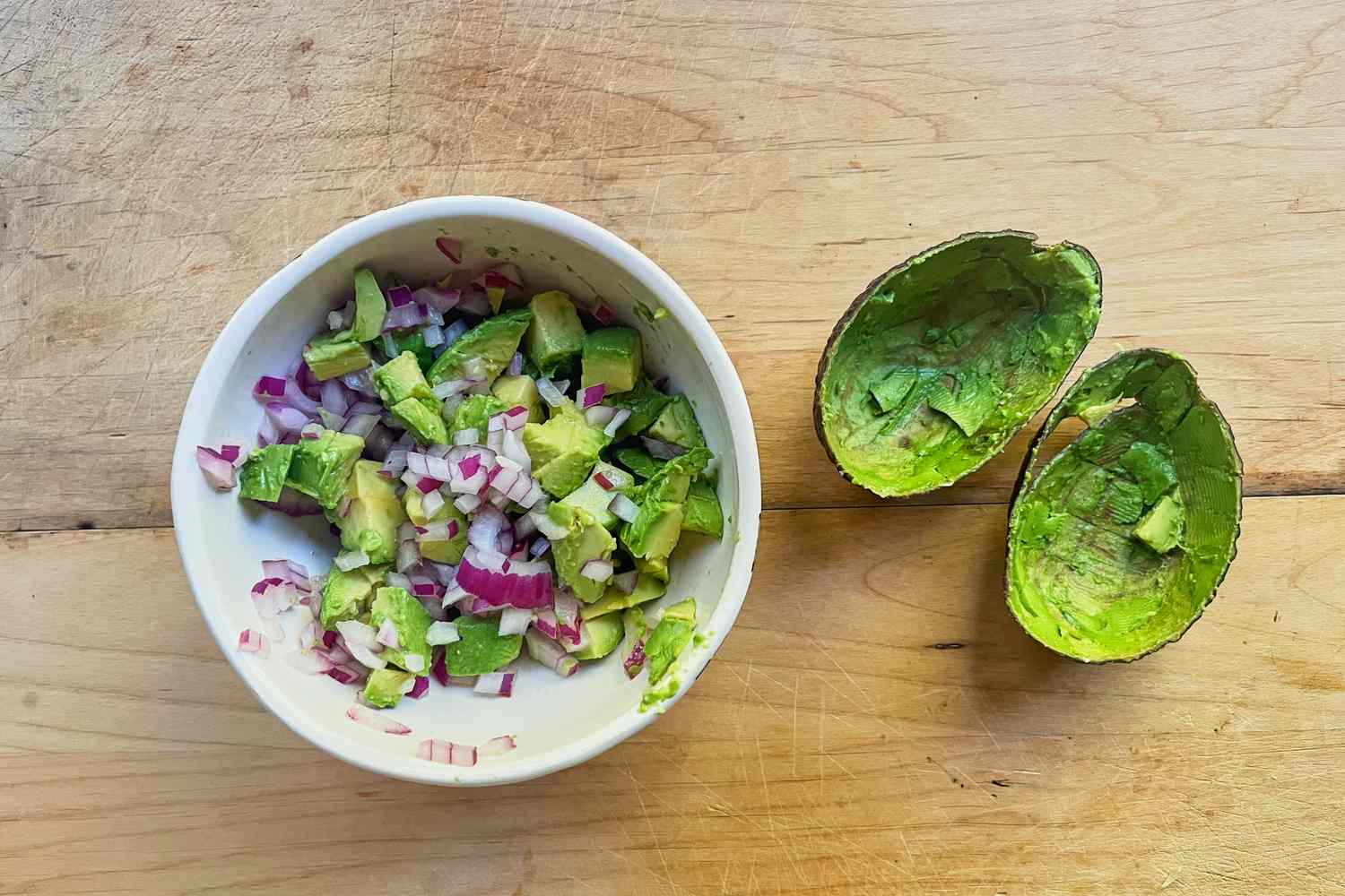 A bowl containing a mixture of diced avocado onion and other ingredients next to emptied avocado skins on a wooden surface