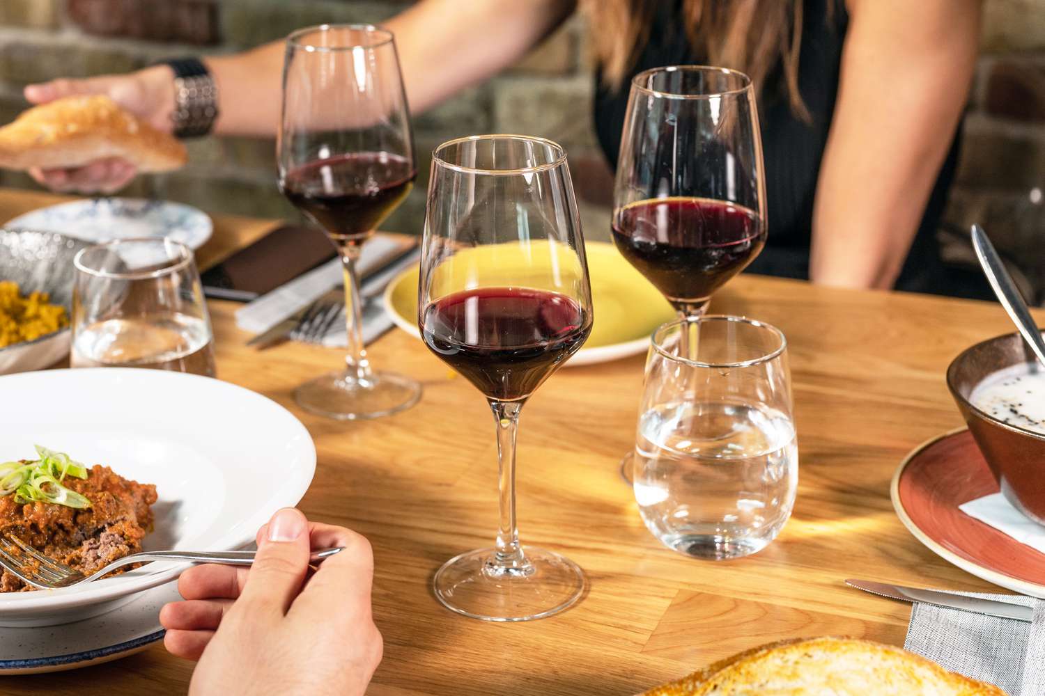 Table settings with people eating dinner and drinking wine. Focus of the photo is the glasses of wine