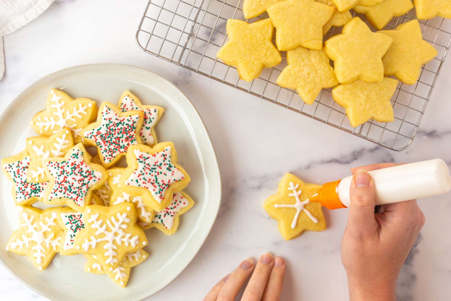A person decorating starshaped cookies with icing a plate and cooling rack of cookies nearby