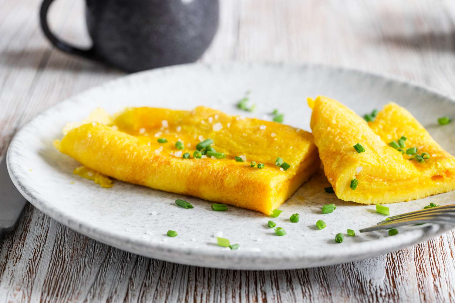 A Frenchstyle omelet garnished with chopped chives on a plate a fork to the side and a mug in the background