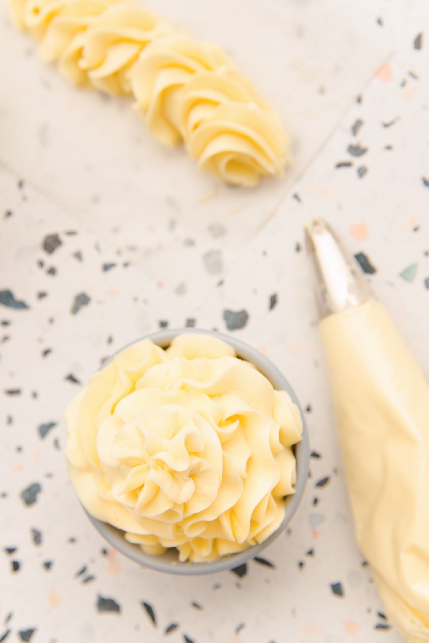 Overhead view of Crème Anglaise frosting in a piping bag and piped on a parchment paper and in a small bowl.