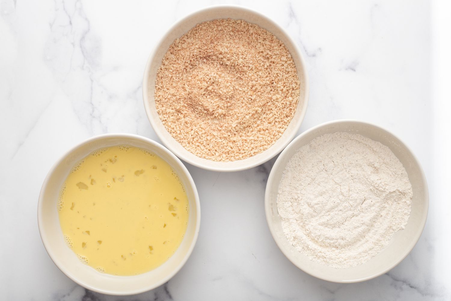 Three bowls filled with breading ingredients to make pork schnitzel.