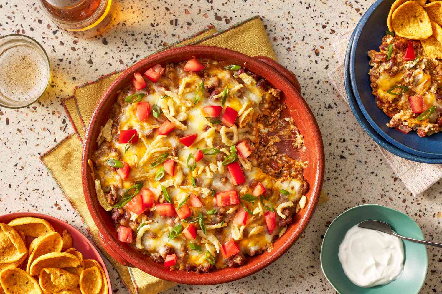 Bowl of chili dip at a table setting with a single serving in a bowl, a bowl with scoop corn chips, a small bowl of cream cheese, and a glasses