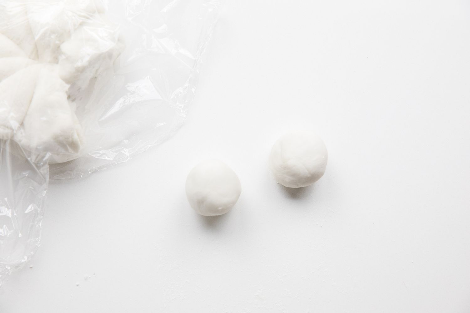 Two Pieces of Dough Rolled into Balls and Placed on the Counter, and to the Right, the Divided Dough Ball Rests under Plastic Wrap 