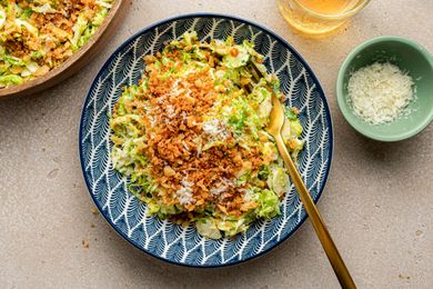 Serving of Brussels Sprouts Caesar Salad with Lemony Breadcrumbs in a Bowl with a Fork Next to a Larger Bowl with More, a Bowl with Shredded Parmesan, and a Glass