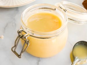 Jar of Homemade Condensed Milk Surrounded by a Spoon With Condensed Milk, a Plate, and a Kitchen Towel