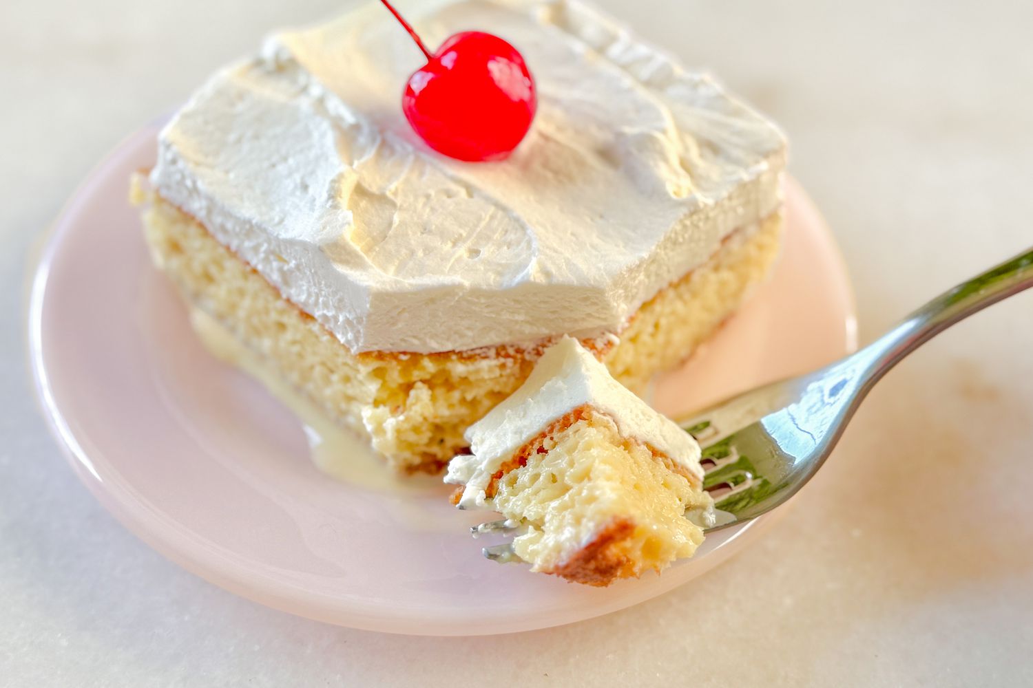 Slice of tres leches cake topped with whipped cream and a cherry, served on a plate with a piece on a fork