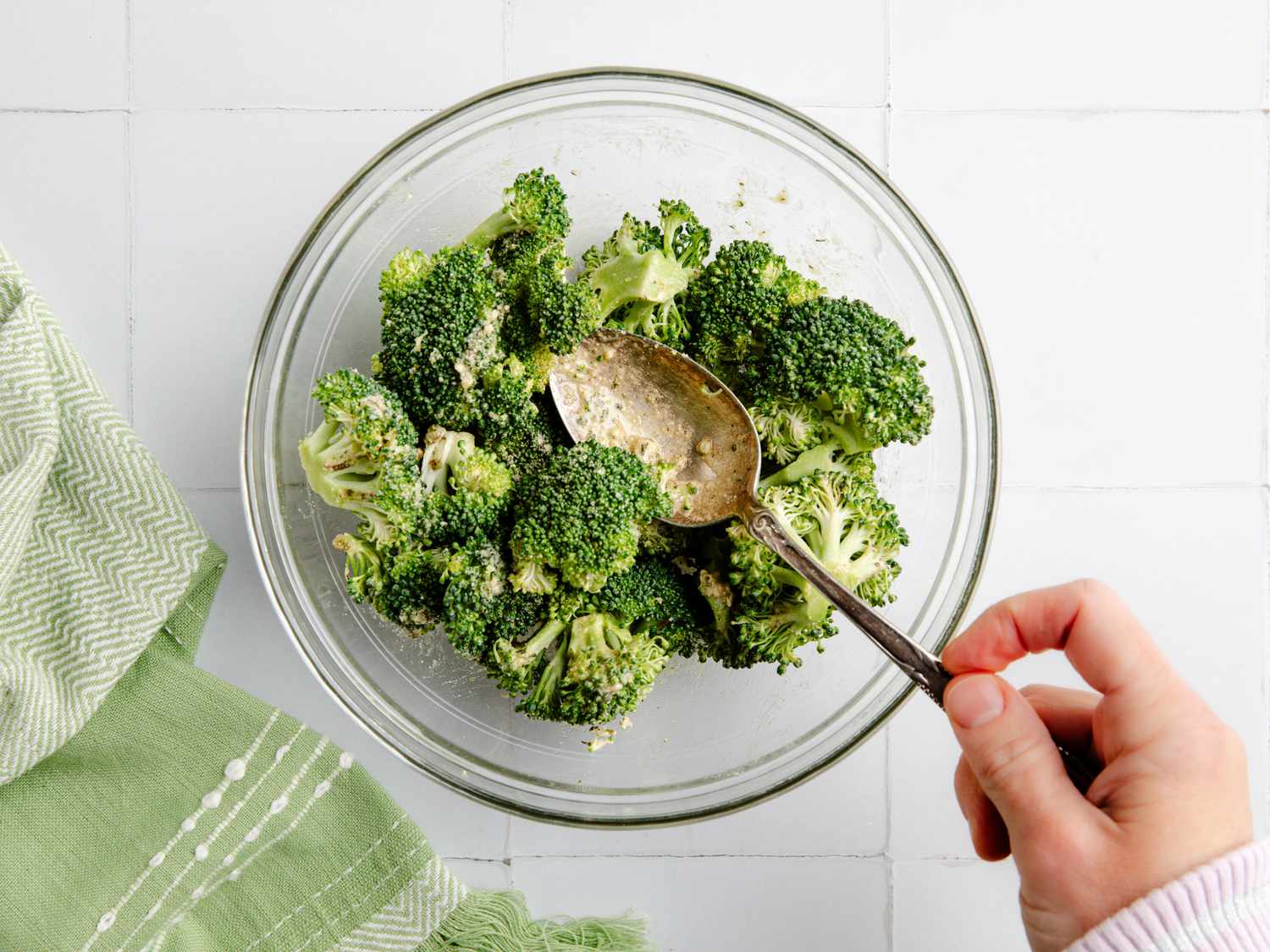 view from above of a hand with a spoon stirring broccoli and oil mix in a bowl 