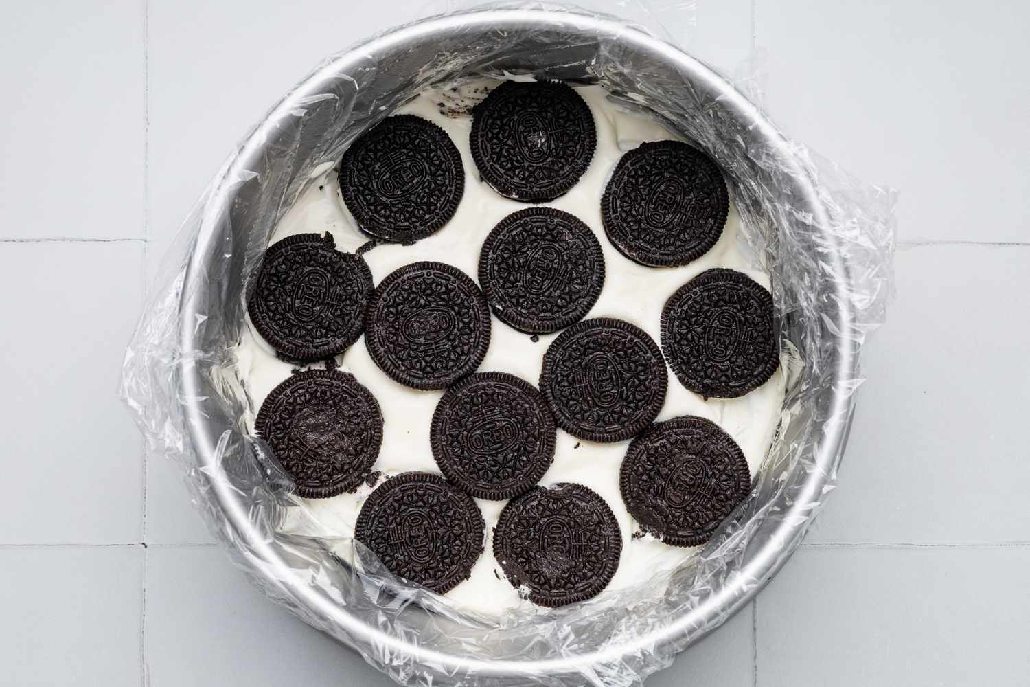 Assembling First Layer of Oreo Ice Cream Cake: Milk Dipped Oreo Cookies on Top of a Layer of No Churn Ice Cream on a Plastic Wrapped Circular Cake Pan