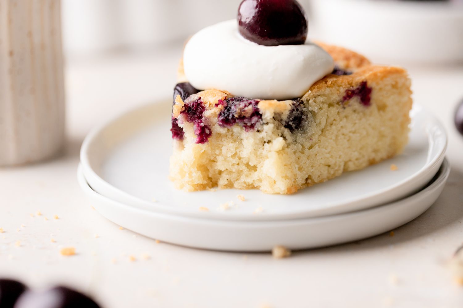 A slice of cherry cake with fresh cherries on a plate and topped with a dollop of whipped cream and a cherry.