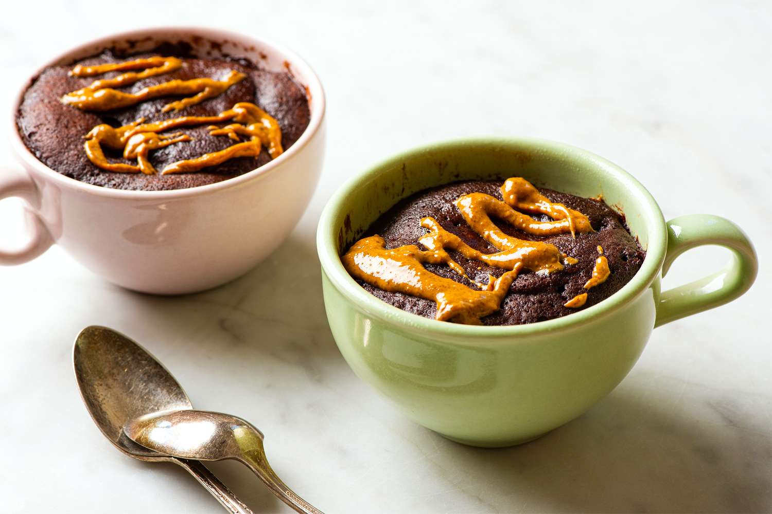 Two mugs of chocolate cake with peanut butter drizzle and two spoons on the side