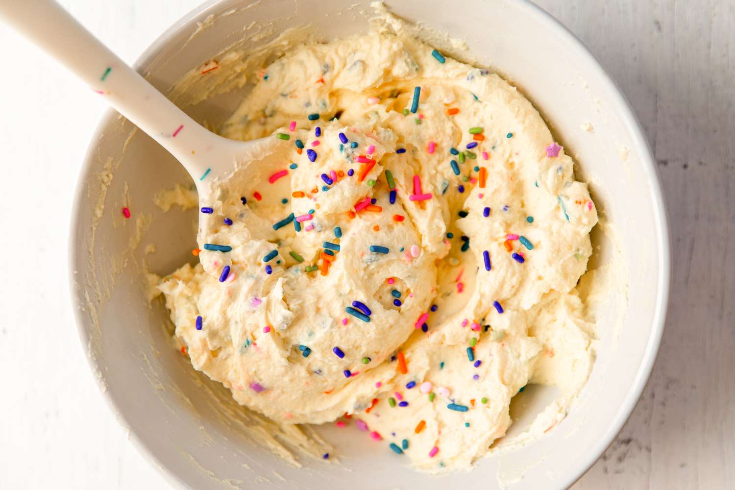 A bowl of yogurt frosting topped with sprinkles, stirred with a spoon