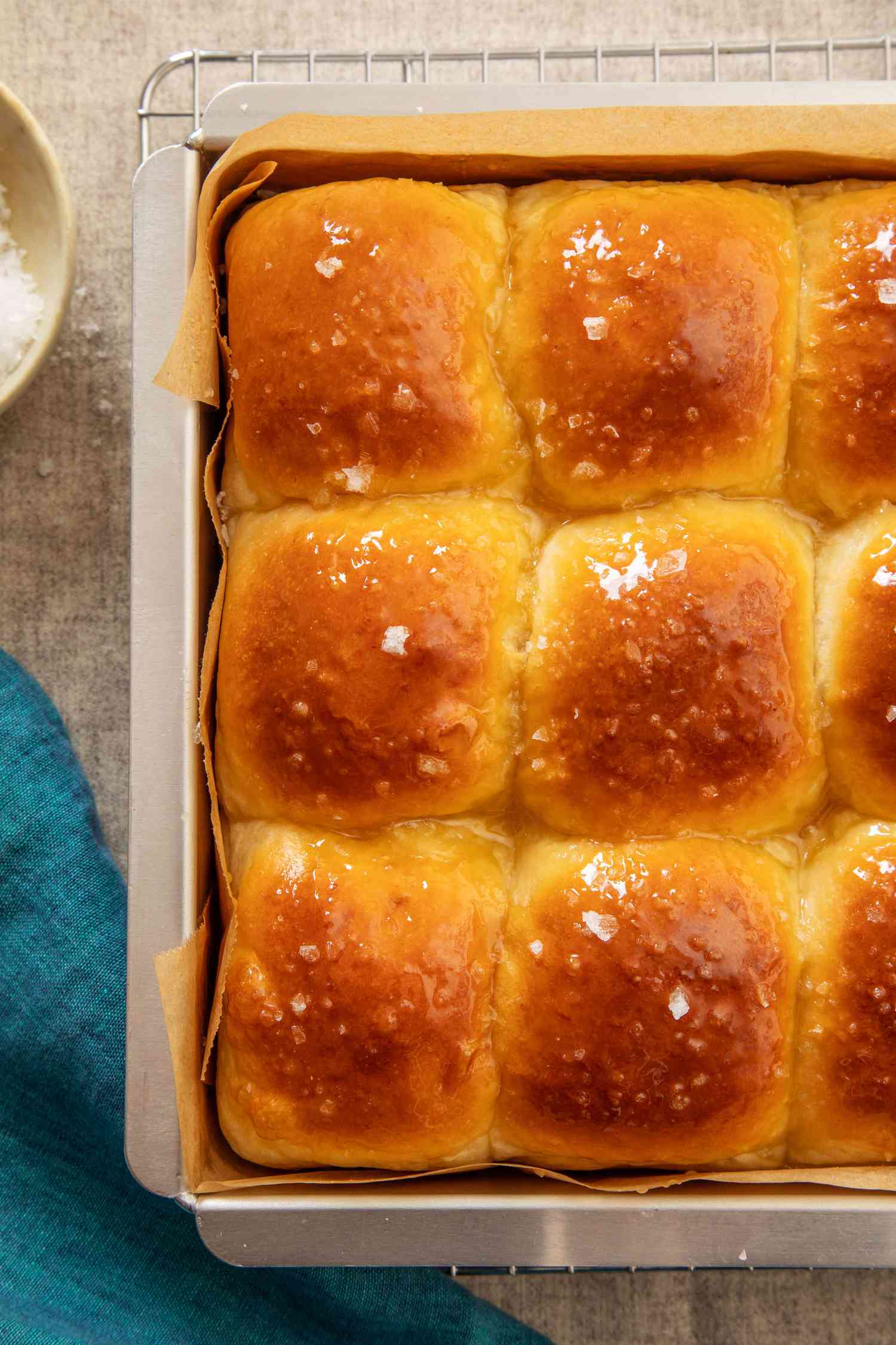 Salted honey rolls in a parchment paper-lined baking sheet on a cooling rack next to a blue kitchen towel
