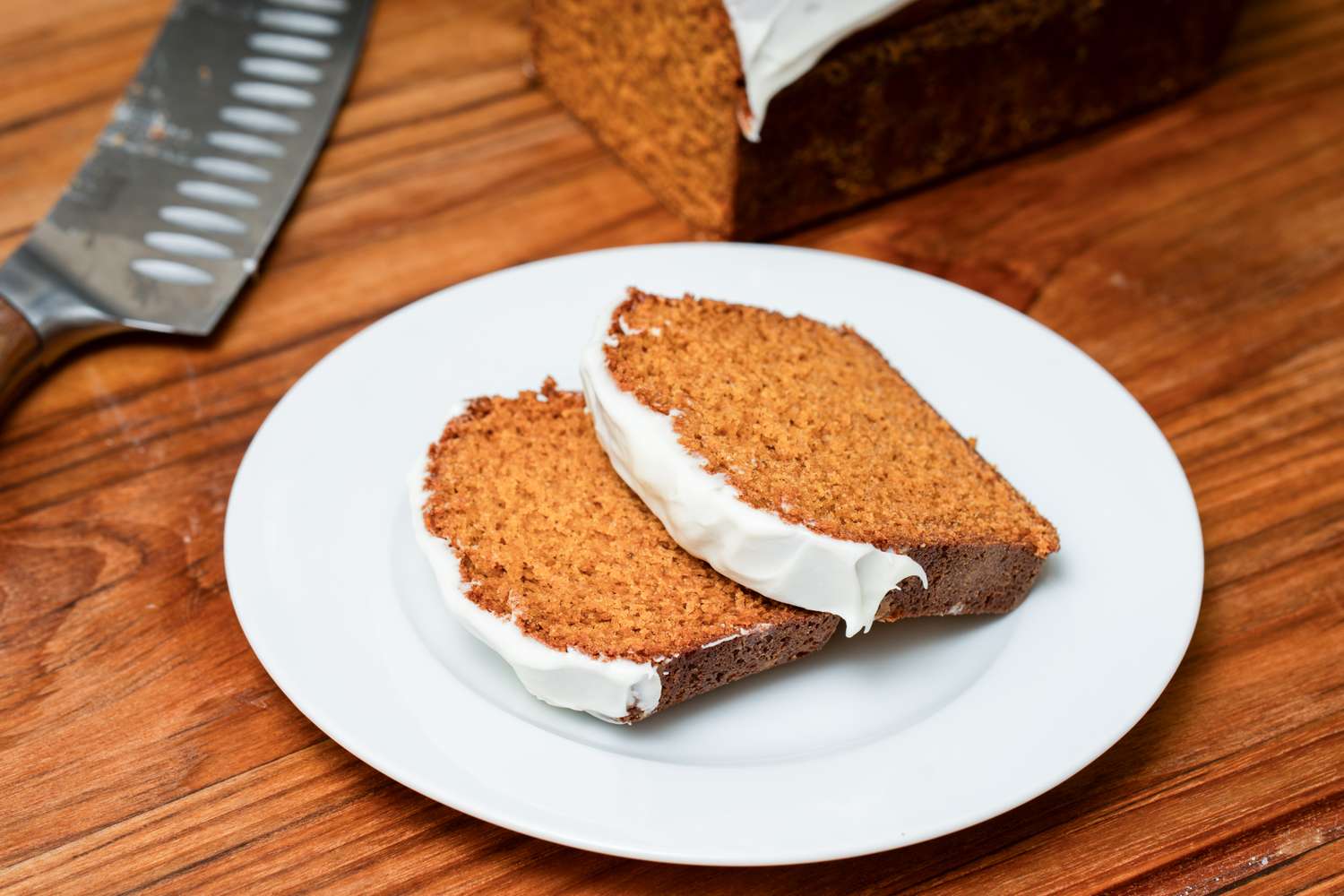 Two slices of frosted loaf cake on a white plate next to a knife on a wooden surface