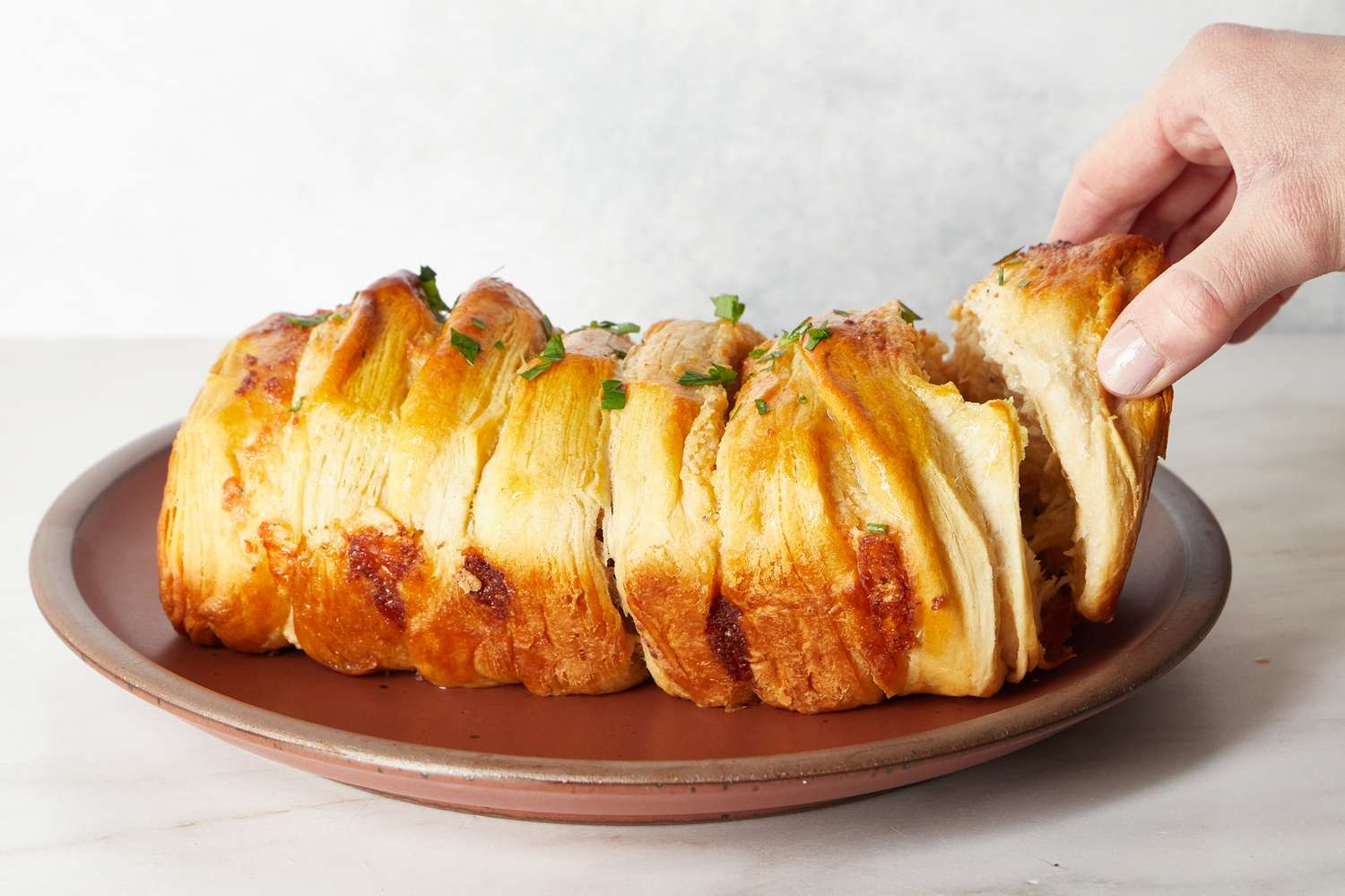 A freshly baked pullapart loaf of Caesarinspired bread on a plate with a hand taking a piece