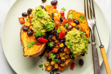 Sweet potato halves topped with black beans corn red peppers guacamole and garnished with cilantro served on a plate with utensils